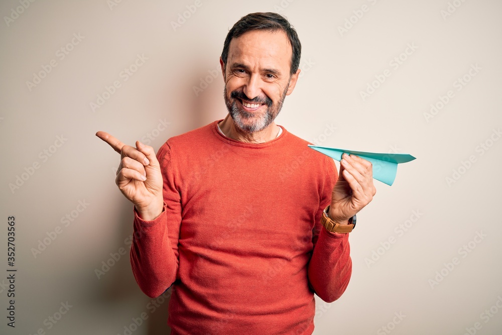 Wall mural Middle age hoary man holding paper aircraft standing over isolated white background very happy pointing with hand and finger to the side