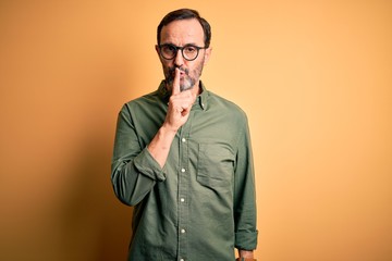 Middle age hoary man wearing casual green shirt and glasses over isolated yellow background asking to be quiet with finger on lips. Silence and secret concept.