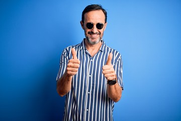 Naklejka premium Middle age hoary man wearing striped shirt and sunglasses over isolated blue background success sign doing positive gesture with hand, thumbs up smiling and happy. Cheerful expression and winner