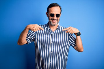 Middle age hoary man wearing striped shirt and sunglasses over isolated blue background looking...