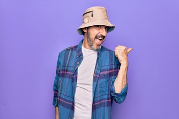 Middle age handsome man wearing casual shirt and hat over isolated purple background pointing thumb up to the side smiling happy with open mouth