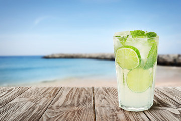 Tasty lemonade with ice cubes and lime on wooden table near sea, space for text