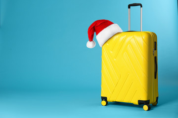 Stylish suitcase with Santa Claus hat on blue background, space for text. Christmas vacation