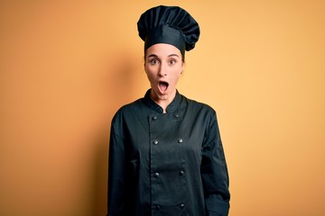 Young beautiful chef woman wearing cooker uniform and hat standing over yellow background afraid...