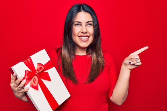 Young beautiful brunette woman holding birthday gift over isolated red background smiling happy pointing with hand and finger to the side