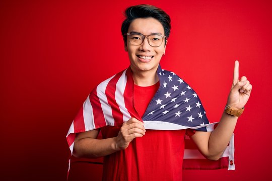 Young chinese patriotic man wearing united states of america flag over red background surprised with an idea or question pointing finger with happy face, number one