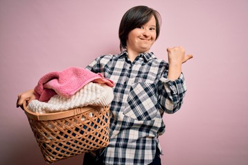 Young down syndrome woman doing housework domestic chores holding laundry wicker basket very happy pointing with hand and finger to the side