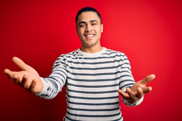 Fototapeta premium Young brazilian man wearing casual striped t-shirt standing over isolated red background smiling cheerful offering hands giving assistance and acceptance.
