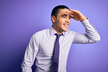 Young brazilian businessman wearing elegant tie standing over isolated purple background very happy and smiling looking far away with hand over head. Searching concept.