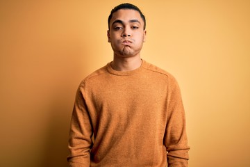 Young brazilian man wearing casual sweater standing over isolated yellow background puffing cheeks with funny face. Mouth inflated with air, crazy expression.