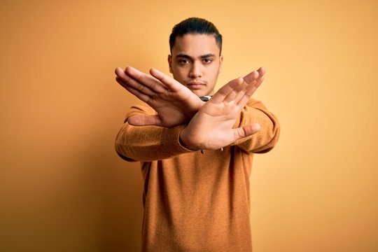 Young brazilian man wearing casual sweater standing over isolated yellow background Rejection expression crossing arms doing negative sign, angry face