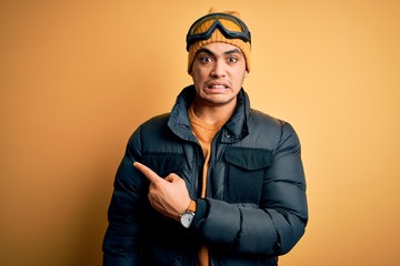 Young brazilian skier man wearing snow sportswear and ski goggles over yellow background Pointing aside worried and nervous with forefinger, concerned and surprised expression