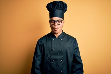 Young brazilian chef man wearing cooker uniform and hat over isolated yellow background making fish face with lips, crazy and comical gesture. Funny expression.