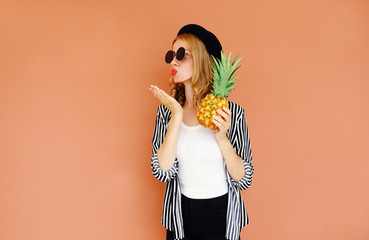 Portrait of stylish woman with pineapple blowing red lips sending sweet air kiss wearing a black hat, sunglasses, striped shirt over wall background