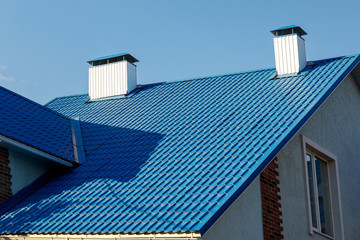 A roof of a house or cottage made of blue metal tiles with drains, slopes and chimney against the blue sky.