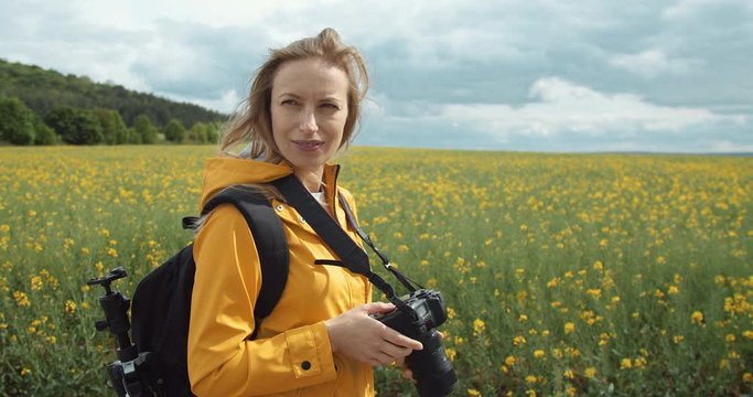 Side view of blonde photographer in yellow jacket with backpack walking through big field and taking pictures on camera. Concept of photography and environment