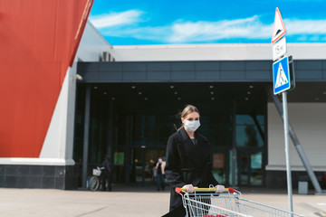 Young woman with a grocery cart on a supermarket background. Social distancing during the Coronavirus Covid-19 pandemic. Disposable mask on face and gloves on hands of a girl. Quarantine shopping