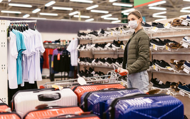 Social distancing concept. A young woman in a disposable mask choosing a suitcase for the upcoming vacation after the end of the Covid-19 coronavirus pandemic. Shopping at supermarket during epidemic