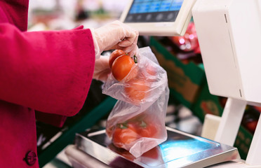 Social distancing in a supermarket. A young woman in a disposable face mask weighing tomatoes and putting them in grocery basket. Shopping during the Coronavirus Covid-19 epidemic 2020