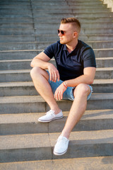 Portrait of attractive young man in sunglasses sitting on stairs being thoughtful in casual wear in the sunset