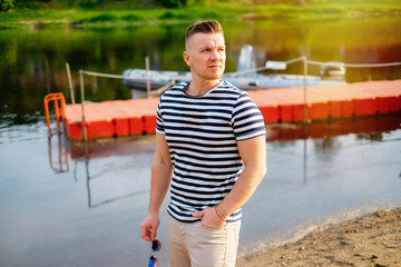 Portrait of attractive young man with sunglasses standing on the beach near the river being thoughtful in casual wear in the sunset