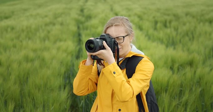 Front view of mature blonde woman in eyeglasses and yellow jacket taking beautiful photos of nature surrounding her. Amateur photographer spending leisure time on green wheat field.