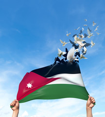 Two hands are holding a Jordan flag which morphs into birds while waving against a blue sky background. 3D Illustration.