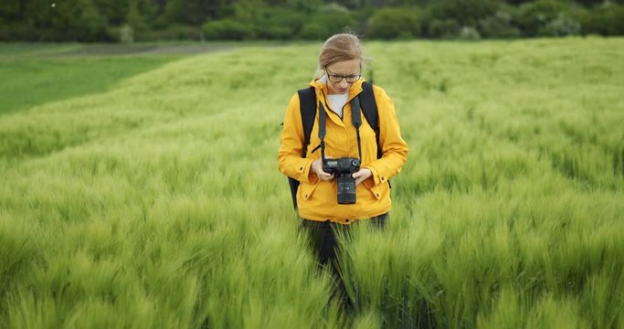 Professional female photographer in casual clothing standing among green wheat field and shooting nature during spring time. Concept of favorite hobby and countryside.
