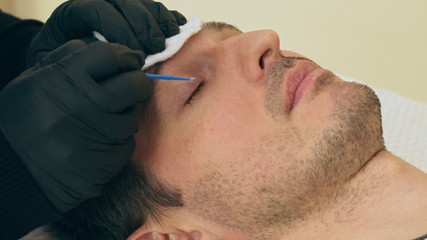 Close up of cosmetologist preparing man for the eyelash extension procedure. High quality close-up of a man laying on a couch during the esthetician lash treatment.