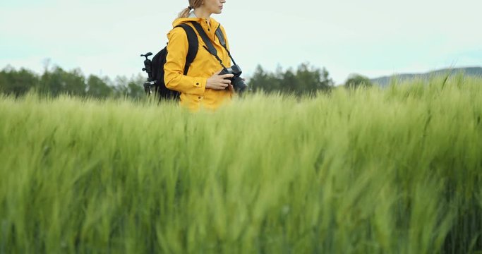 Side view of beautiful woman in yellow jacket spending free time for shooting green wheat field. Female photographer with backpack enjoying spring nature around.