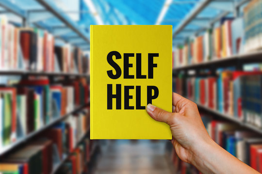 A hand holding 'Self-Help' book in a library.