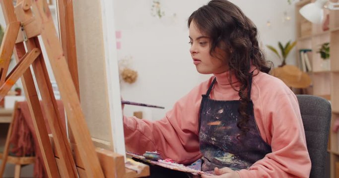 Pretty girl with down syndrome painting and singing in her room. Concentrated chid wearing aprone holding art palette and soaking brush into paint while sitting in front of molbert.