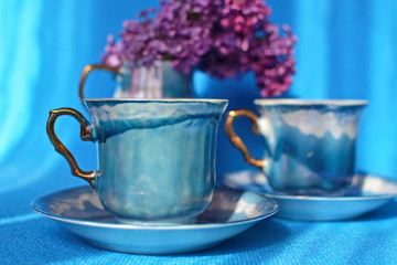 Beautiful couple blue porcelain cup of tea with golden handle on saucer on blurred background of cermic vase with blooming bunch of purple lilac. Selective focus. Horizontal frame