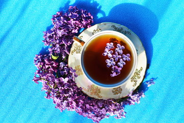 Blooming branch of purple lilac around pattern saucer with porcelain Cup of brown tea with flower...