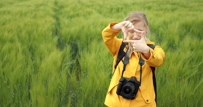 Beautiful woman in yellow jacket and black trousers making frame with fingers while standing among green wheat field. Female photographer looking for perfect composition for shooting outdoors.