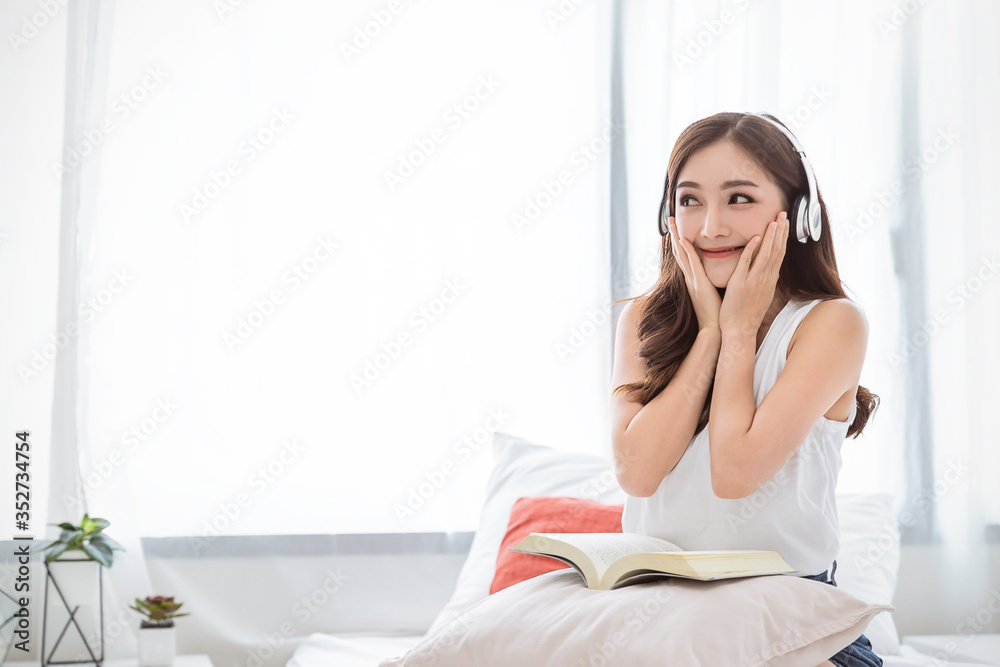 Wall mural Portrait of young beautiful asian woman relax holding book listen to music from headphone in bedroom. Smile happy asian girl relaxing. Knowledge education university lifestyle, back to school concept