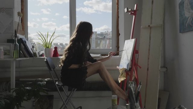 long haired girl with slim legs in short black dress draws picture sitting at easel by window in art studio at home slow motion