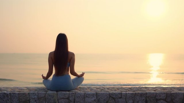Back to camera, A young woman sits meditating on a stone wall facing the incoming tide at sunset. copy space