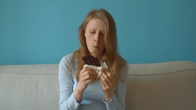 Young beautiful woman sitting on the couch in the room and eating chocolate. Contravention of diet, enjoyment of life, hedonism