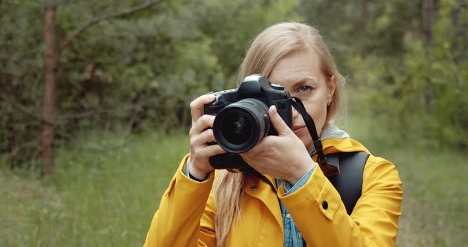 Close up of concentrated mature lady with blond hair using professional camera for taking pictures of spring forest. Concept of working process outdoors.