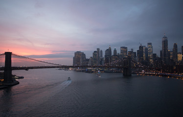Naklejka premium New York City, USA - 26 Dec 2019: Dramatic, colorful Sunset at the East River with Manhattan Skyline Lights and Brooklyn Bridge