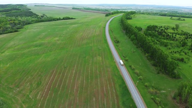 A lorry with a white semi-trailer is driving along the highway surrounded by a green field. Quadrocopter aerial photography