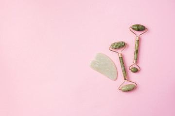 Green jade face roller and gua-sha scraper for facial acupuncture massage on pink background. Copy...