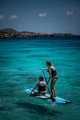 2 girls on a paddle board while wearing bikinis and lifejackets. Blue open ocean and blue sky
