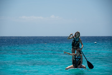 2 girls using a stand up paddle board on the open blue ocean while wearing bikinis and life jackets. One sits and one uses the paddle