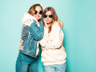 Two young beautiful brunette smiling hipster girls in trendy summer hoodie and jeans jacket clothes. Sexy carefree women posing near blue wall. Trendy and positive models having fun in sunglasses