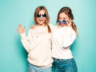 Two young beautiful blond smiling hipster girls in trendy summer hoodie clothes. Sexy carefree women posing near blue wall. Trendy and positive models having fun in sunglasses