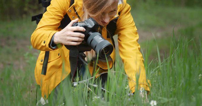 Close up of professional photographer in yellow jacket taking pictures of green forest, using tripod and digital camera. Mature woman with blond hair enjoying favorite hobby on fresh air.