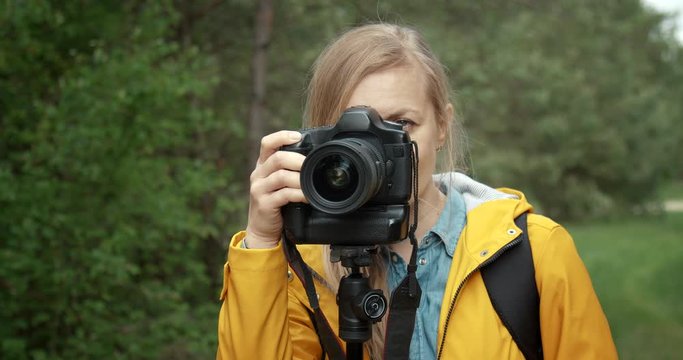 Close up of blonde photographer in yellow jacket standing near tripod with digital camera and taking pictures of wild nature at spring forest. Concept of adventure and environment