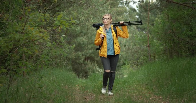 Full length portrait of mature lady in glasses, yellow jacket and black trousers walking through green forest. Competent photographer carrying on big backpack and tripod for shooting outdoors.